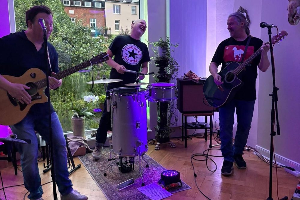 Plans For Nigel bei einem Wohnzimmerkonzert – drei Musiker mit Akustikgitarren und Schlagzeug in stimmungsvollem Licht